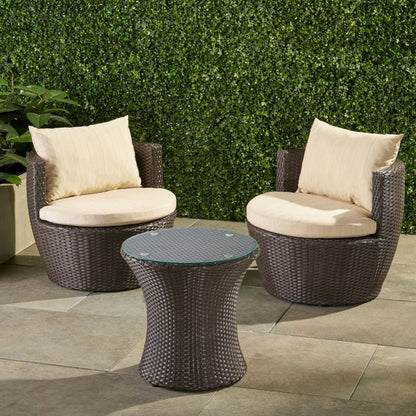 The Kono - Chat Set Outdoor Furniture features two round wicker patio chairs with beige cushions and a matching wicker side table with a glass top, arranged on a tiled patio with a green hedge in the background.