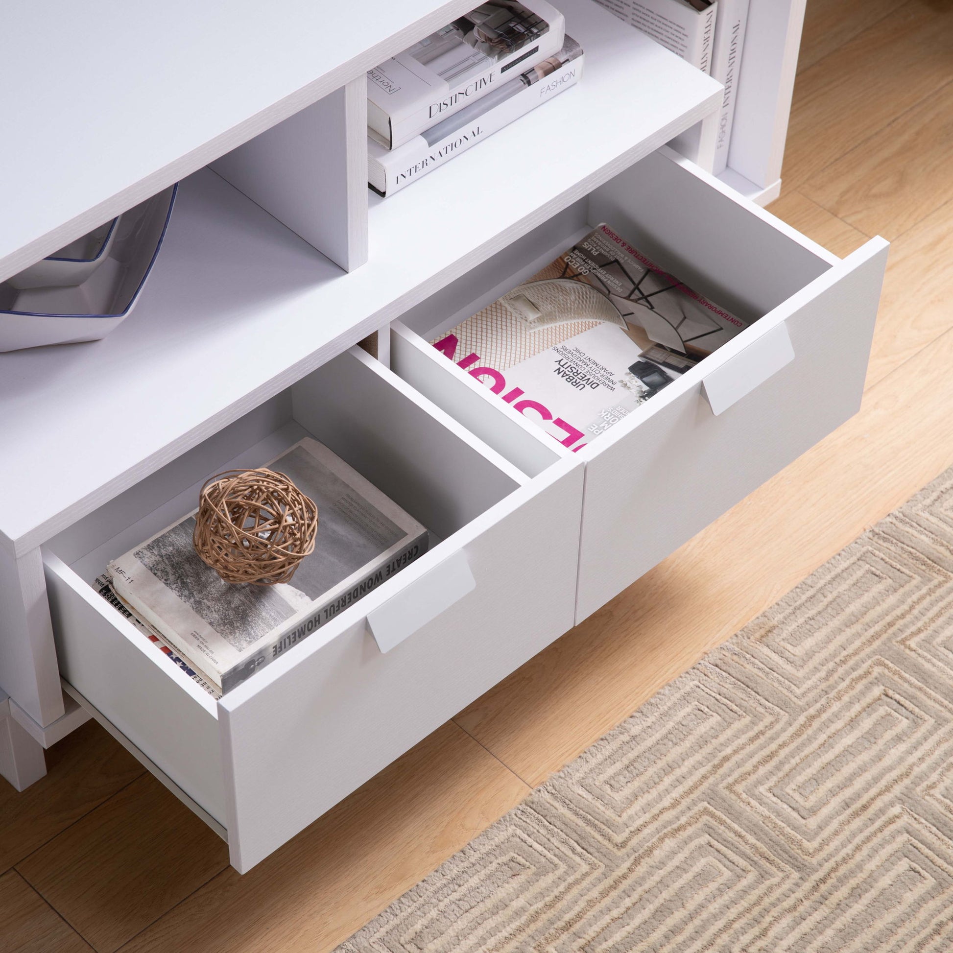 The Modern TV Stand features two open white drawers holding magazines, books, and a decorative woven ball, set on a light wood floor beside a beige patterned rug.