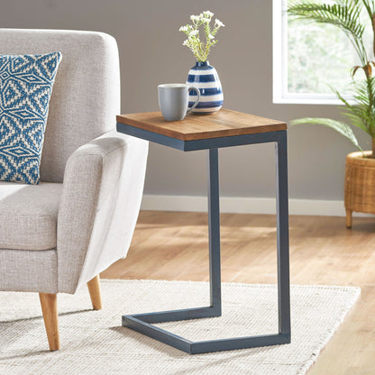 The C Shaped End Table With Iron Base And Fir Wood Top stands next to a light gray sofa, holding a striped vase with flowers and a mug. A cushion, potted plant, and bright window appear in the background.