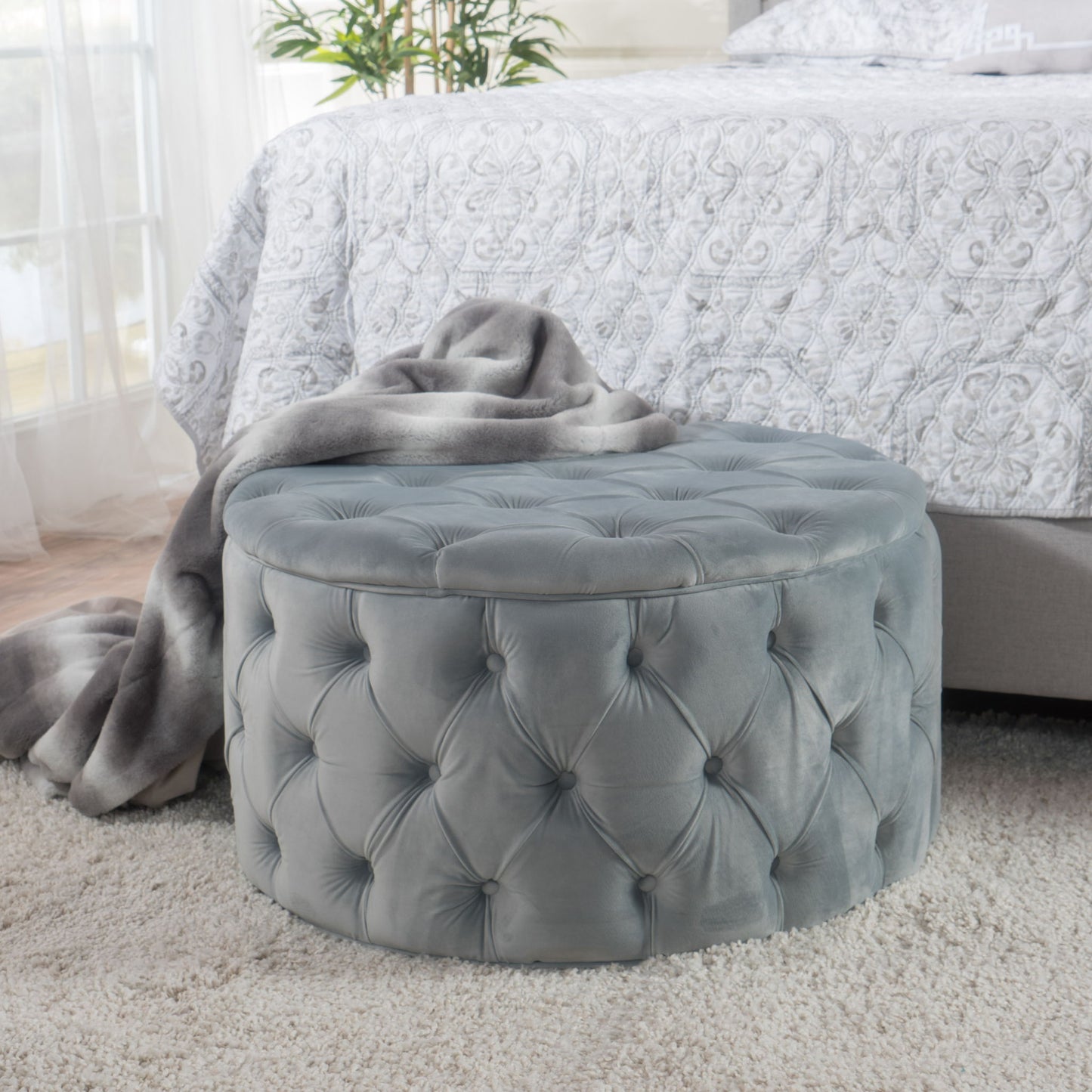 A Modern Glam Ottoman Velvet in gray sits on a plush light carpet by a bed with a patterned white comforter, draped with a gray blanket. A leafy plant is visible in the background.