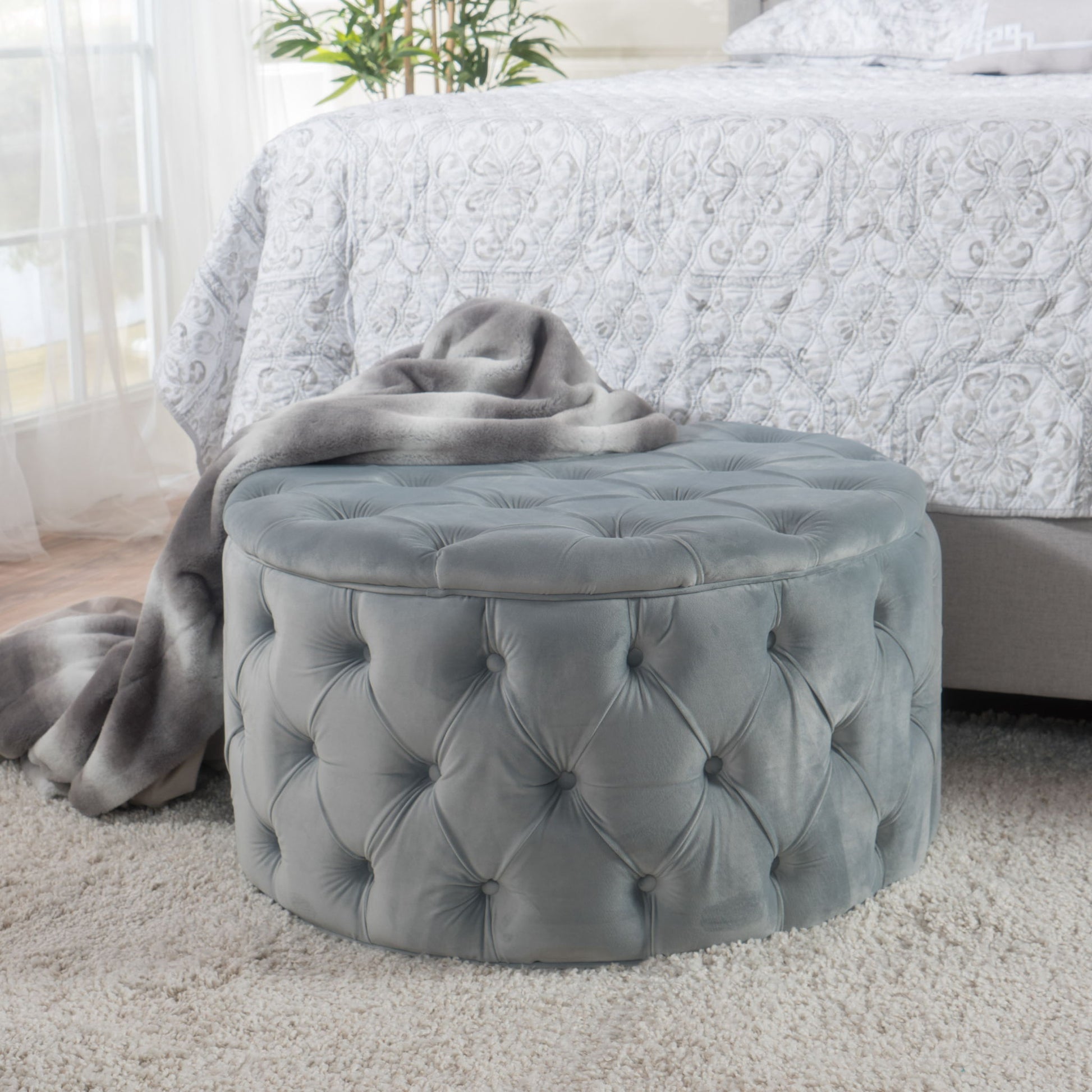 A Modern Glam Ottoman Velvet in gray sits on a plush light carpet by a bed with a patterned white comforter, draped with a gray blanket. A leafy plant is visible in the background.