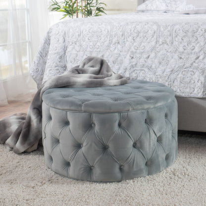 A Modern Glam Ottoman Velvet in gray sits on a plush light carpet by a bed with a patterned white comforter, draped with a gray blanket. A leafy plant is visible in the background.