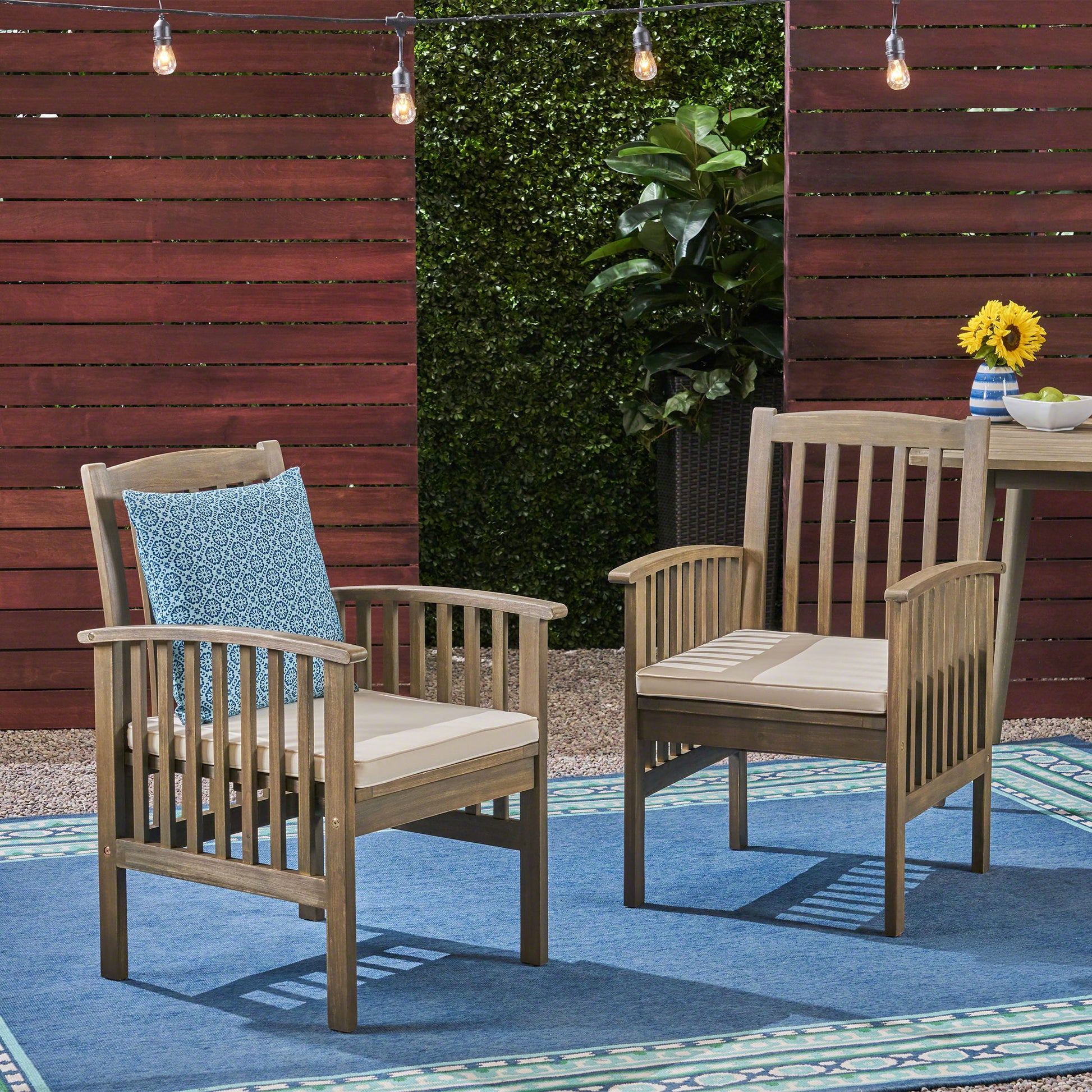 Two Casa Acacia Dining Chairs with beige cushions are on a blue outdoor rug. One chair features a blue patterned pillow. Behind them are a red fence, string lights, green hedge, and a small table topped with sunflowers.