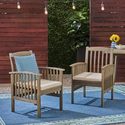 Two Casa Acacia Dining Chairs with beige cushions are on a blue outdoor rug. One chair features a blue patterned pillow. Behind them are a red fence, string lights, green hedge, and a small table topped with sunflowers.