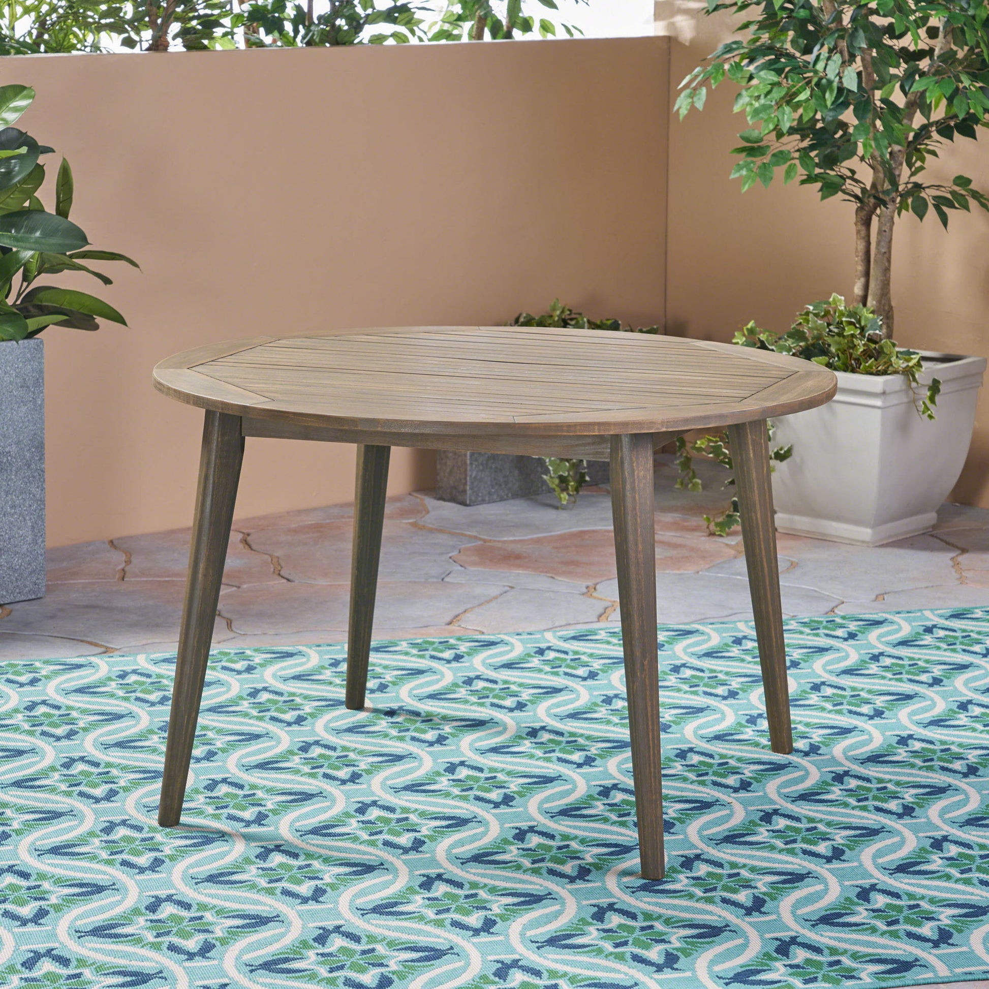 The Stamford - Outdoor Dining Table Modern Design, featuring a round wooden top and four angled legs, sits on a blue and white patterned outdoor rug and is surrounded by green potted plants on a stone patio.