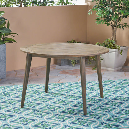 The Stamford - Outdoor Dining Table Modern Design, featuring a round wooden top and four angled legs, sits on a blue and white patterned outdoor rug and is surrounded by green potted plants on a stone patio.