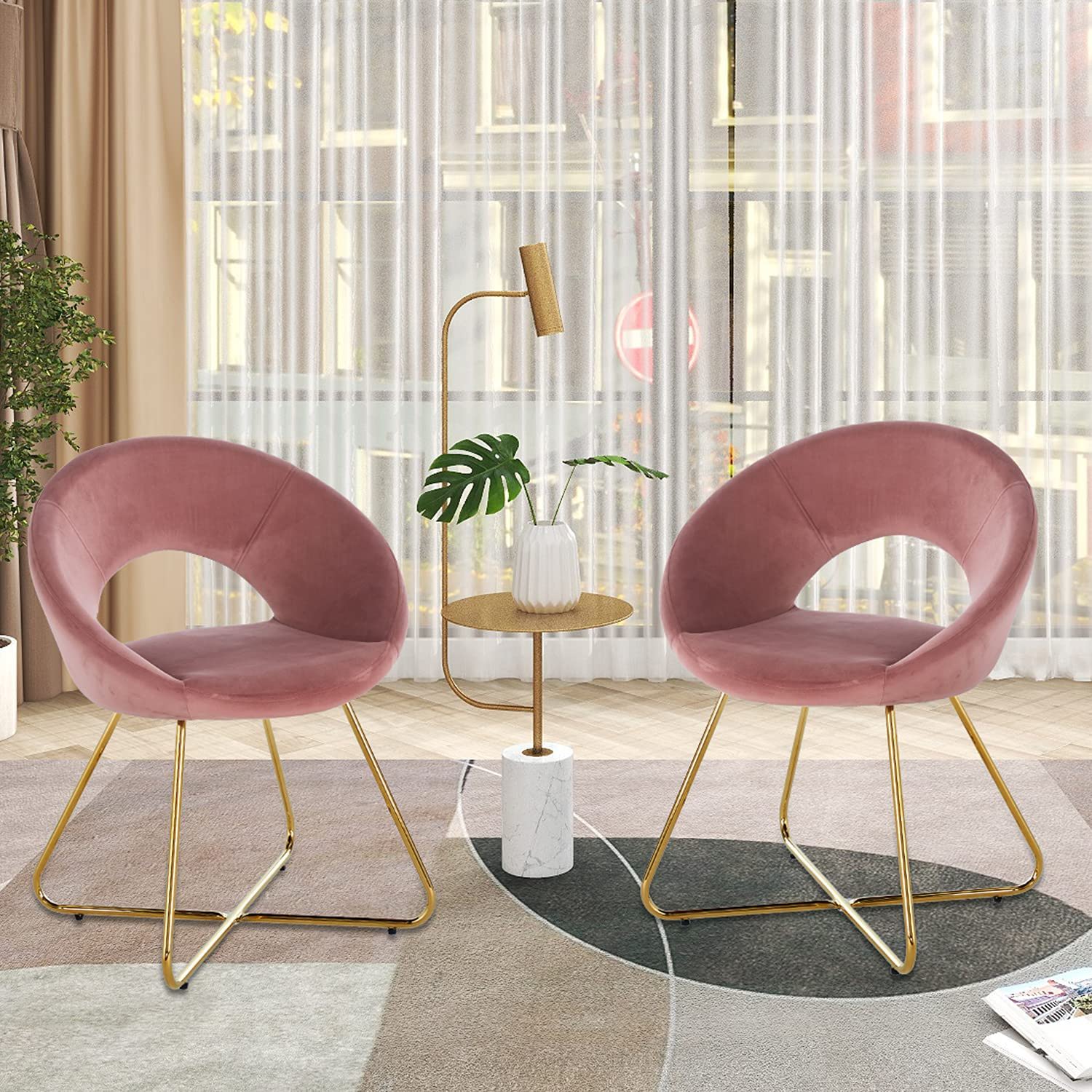 Two Modern Accent Velvet Lounge Chairs with gold legs face each other across a small round table with a marble base, decorative leaf in vase, and gold floor lamp in a bright, stylish living room with large windows.