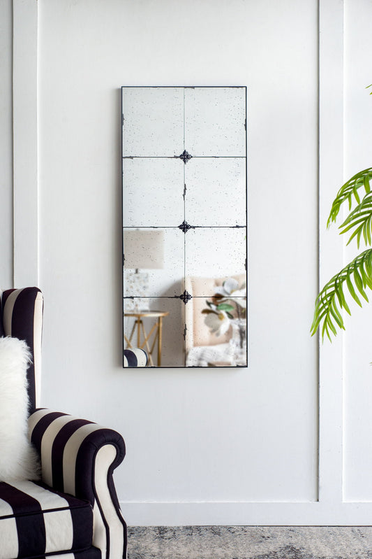 The Traditional Decor Style Antique Glass Panel Mirror hangs on a white wall, flanked by a black-and-white striped armchair and a leafy green plant. Its vintage segmented design reflects a blurred, elegant interior.