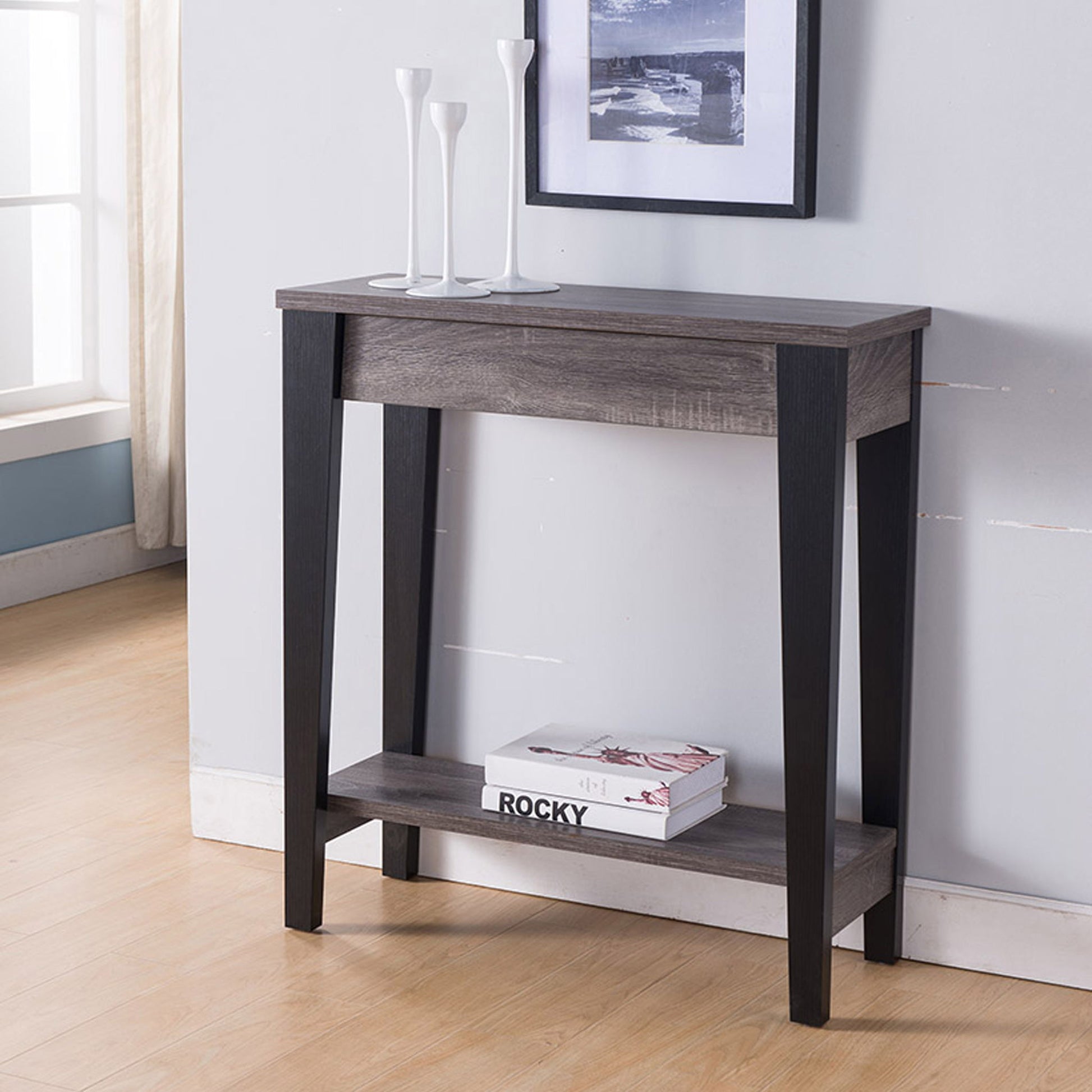 The Modern Two Toned Console With Two Shelves, featuring black legs and a lower shelf with books, is topped with three white vases and placed against a wall beneath a framed picture in a bright room.