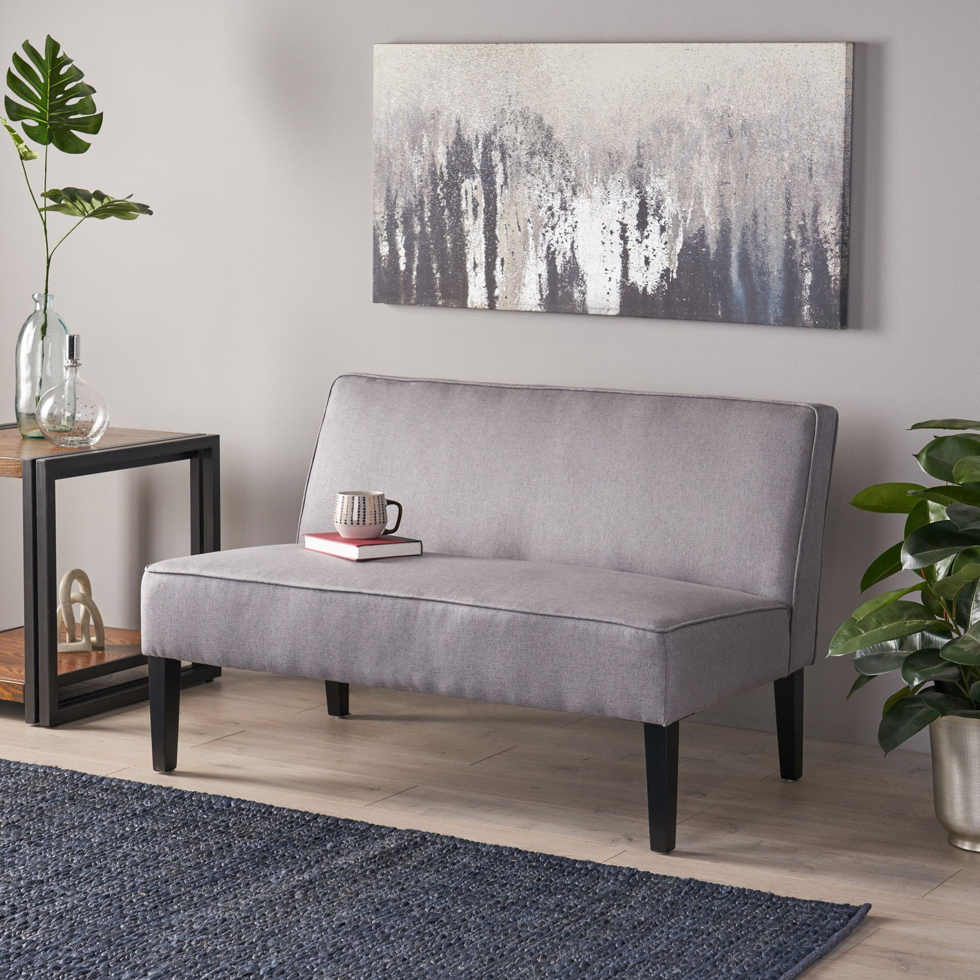 A modern living room featuring a Contemporary Loveseat With Upholstered Fabric And Birch Wood Legs in light gray, a blue rug, side table with decor, potted plants, abstract gray and white wall art, plus a mug and book on the loveseat.
