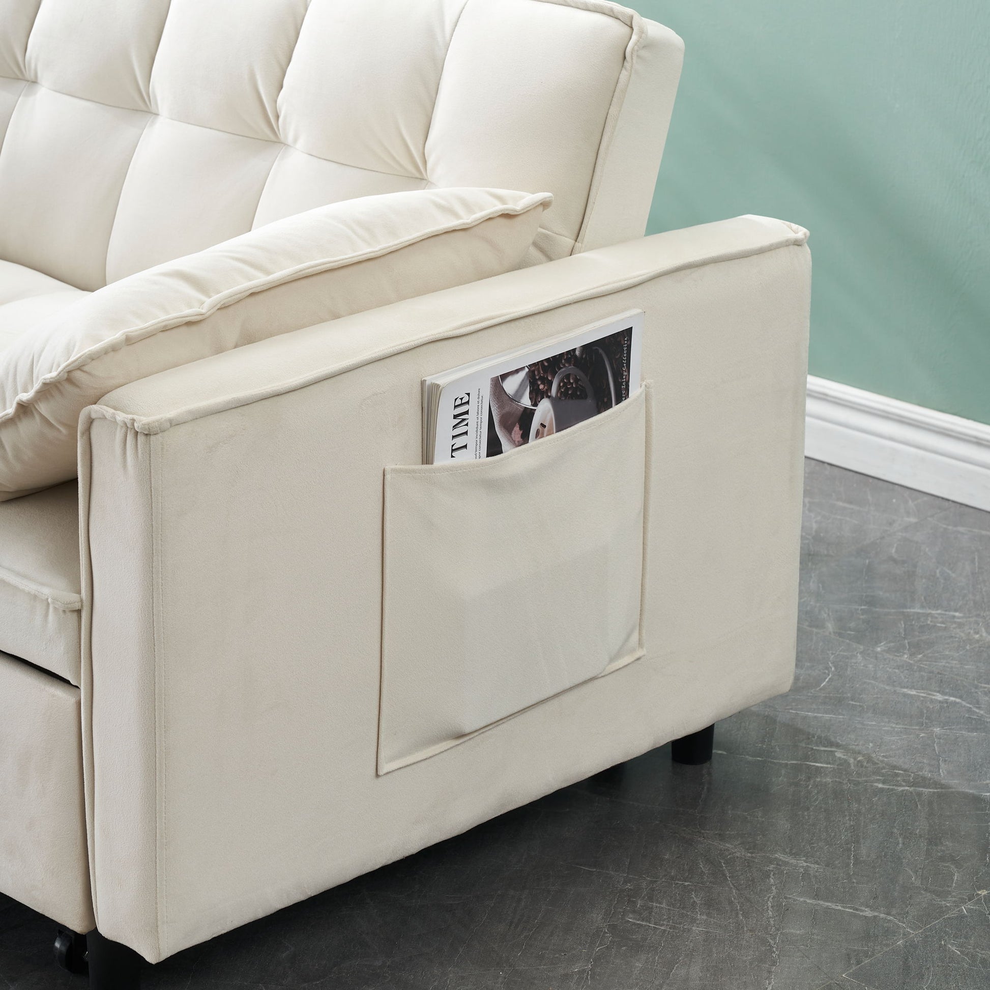 The Modern Velvet Sofa features a pull-out bed, cushioned armrest, side pocket for magazines, and includes a pillow. This 3-in-1 convertible sleeper sofa sits on a dark textured floor against a light green wall with white trim.