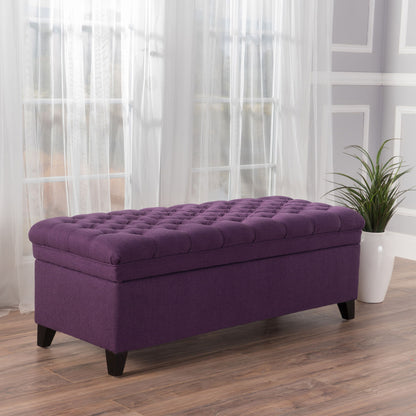 The Juliana Storage Ottoman with birch legs sits on a wooden floor in front of sheer curtains, with a potted plant nearby and gray paneled walls in the background.