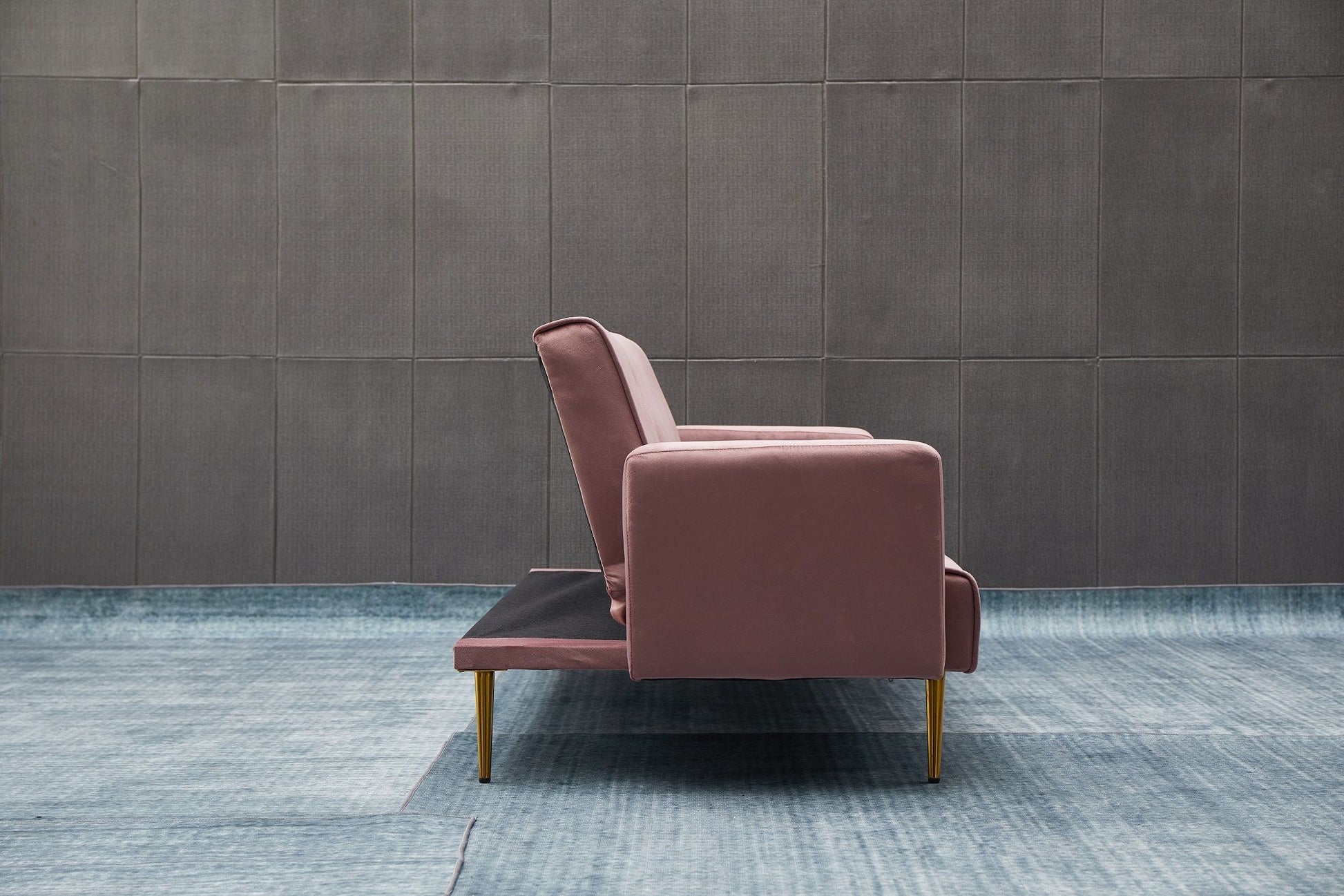 The Comfort Compact Design Sofa Bed, featuring a modern pink look with gold legs, is shown from the side on a light blue textured floor in front of a dark gray tiled wall.