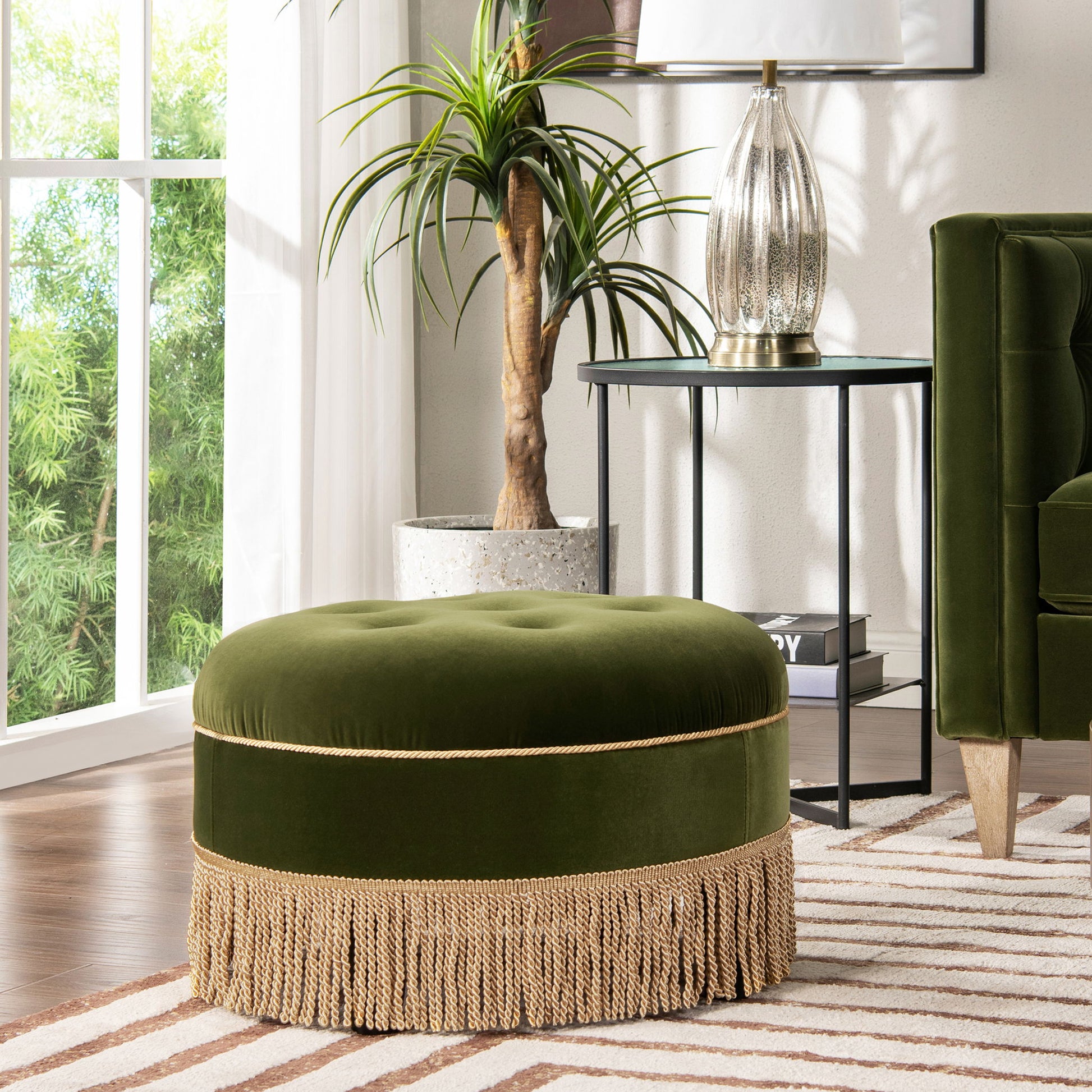 The Yolanda Round Upholstered Accent Ottoman, in green velvet with gold fringe, sits on a striped rug by a green armchair, potted plant, and side table with decor in a sunlit living room.