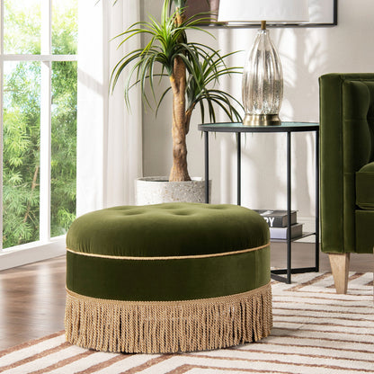 The Yolanda Round Upholstered Accent Ottoman, in green velvet with gold fringe, sits on a striped rug by a green armchair, potted plant, and side table with decor in a sunlit living room.