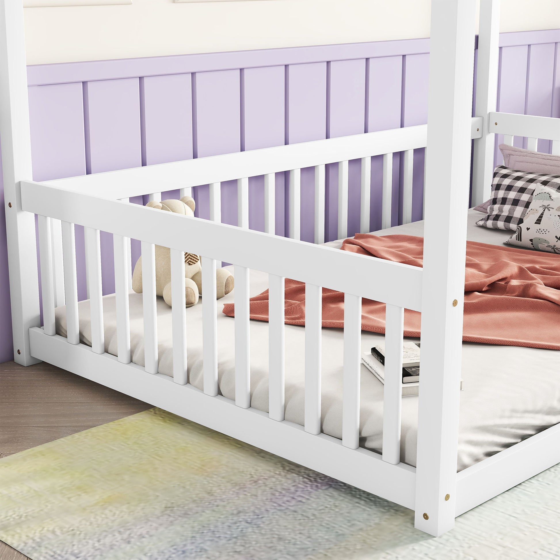 The Canopy Frame Floor Bed With Fence, Guardrails features white wooden rails and sits against a purple paneled wall. On the bed are a teddy bear, checkered pillows, and a pink blanket, with a soft rug placed on the floor nearby.