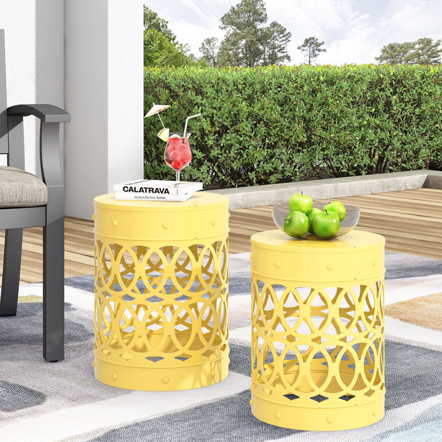 Two Outdoor Metal S/2 End Tables with circular cut-outs are placed on a patio rug. One holds green apples in a glass bowl, the other displays a stack of books and a red cocktail. Green bushes and trees form the background.