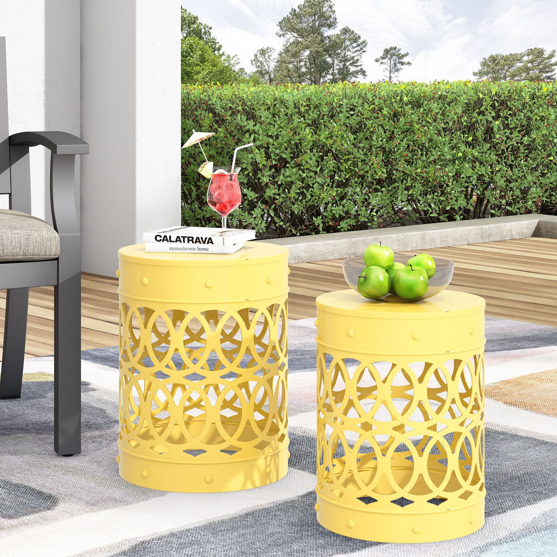 Two Outdoor Metal S/2 End Tables with circular cut-outs are placed on a patio rug. One holds green apples in a glass bowl, the other displays a stack of books and a red cocktail. Green bushes and trees form the background.