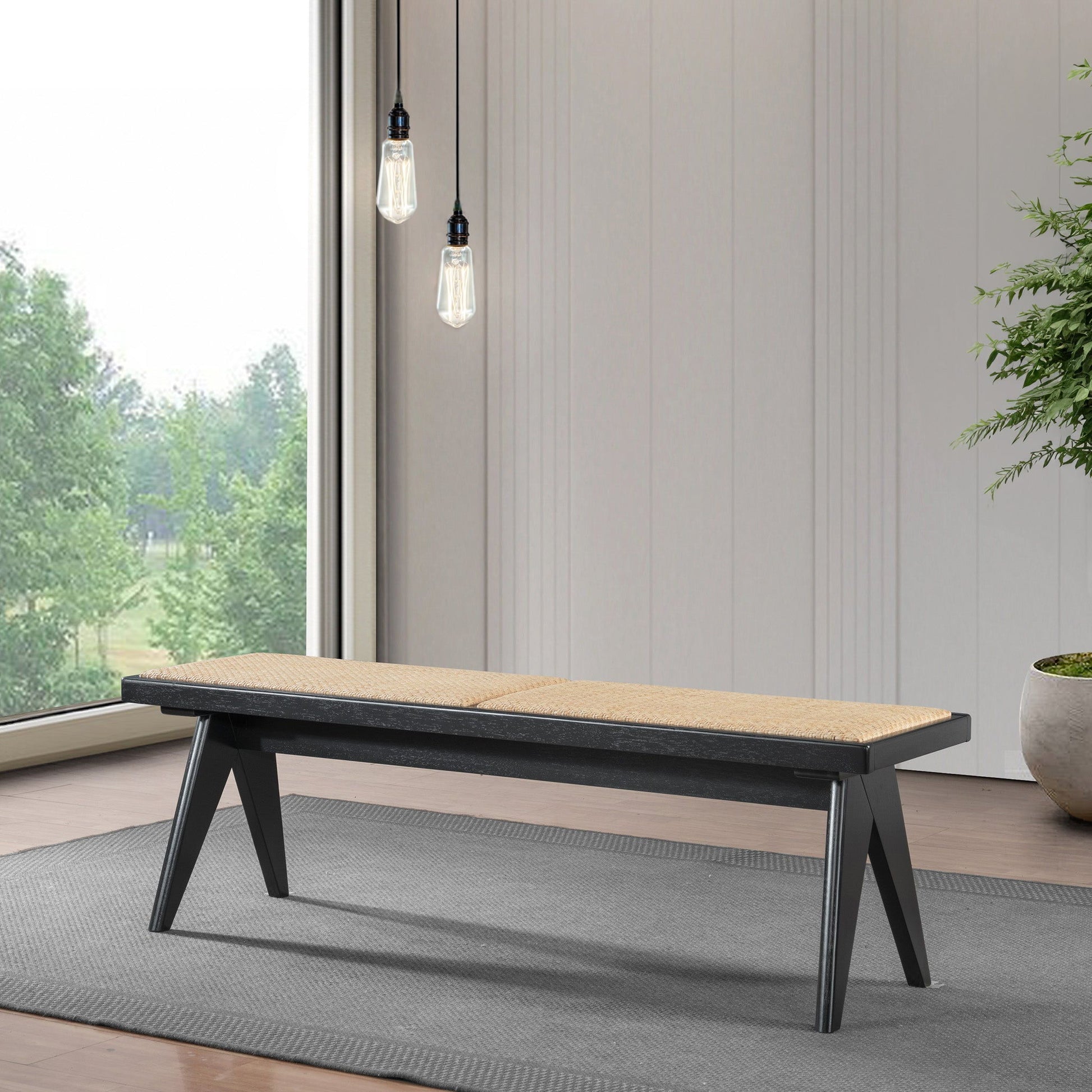 The Keira - Modern Aesthetics Bench, featuring a light tan cushioned seat and black angled legs, rests on a gray rug in a minimalist room with large windows overlooking trees and two hanging light bulbs in the background.