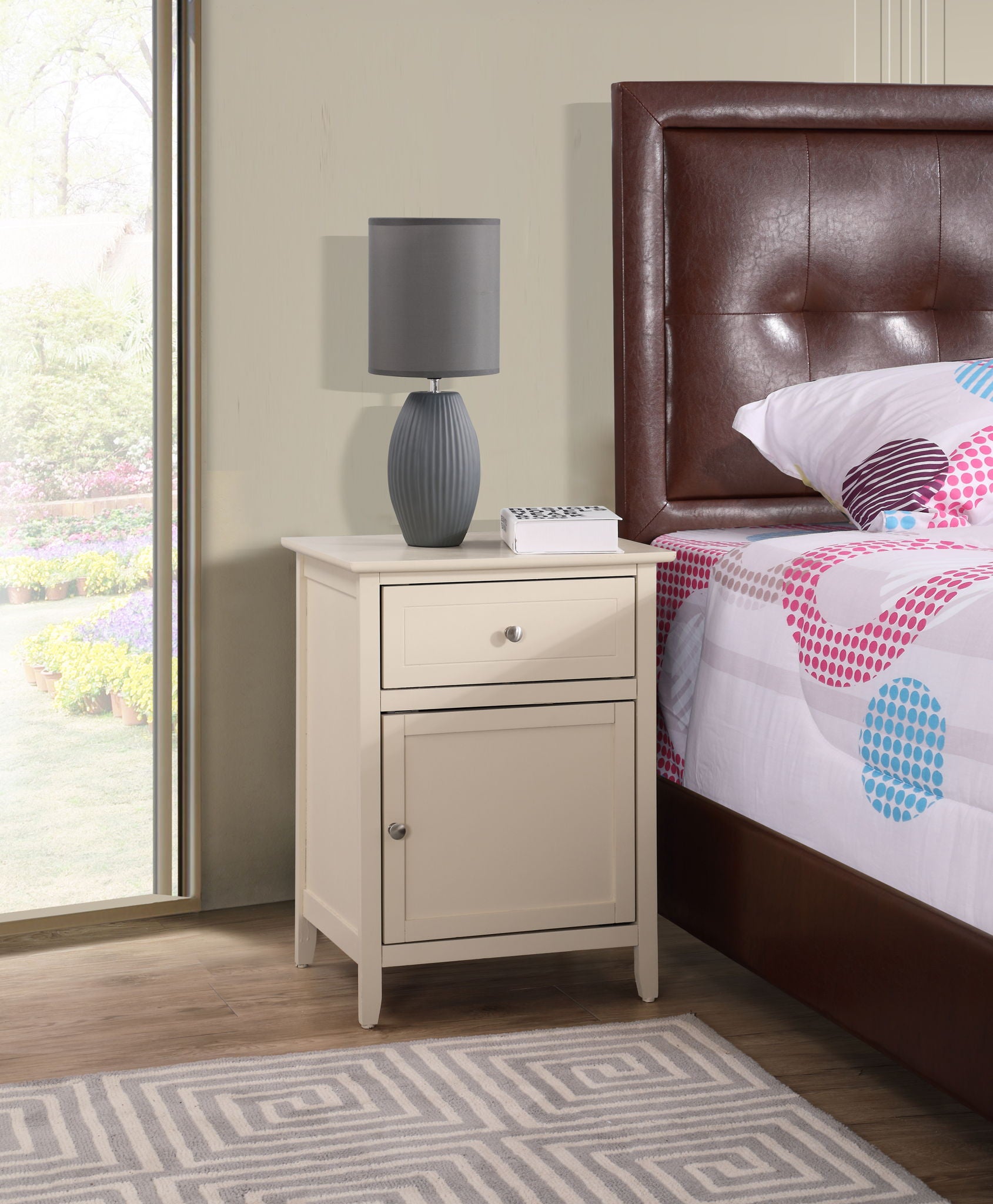 The Izzy - 1 Drawer / 1 Door Nightstand in cream stands by a bed with a brown upholstered headboard. A gray table lamp and a book rest on the nightstand, next to a window overlooking the garden.