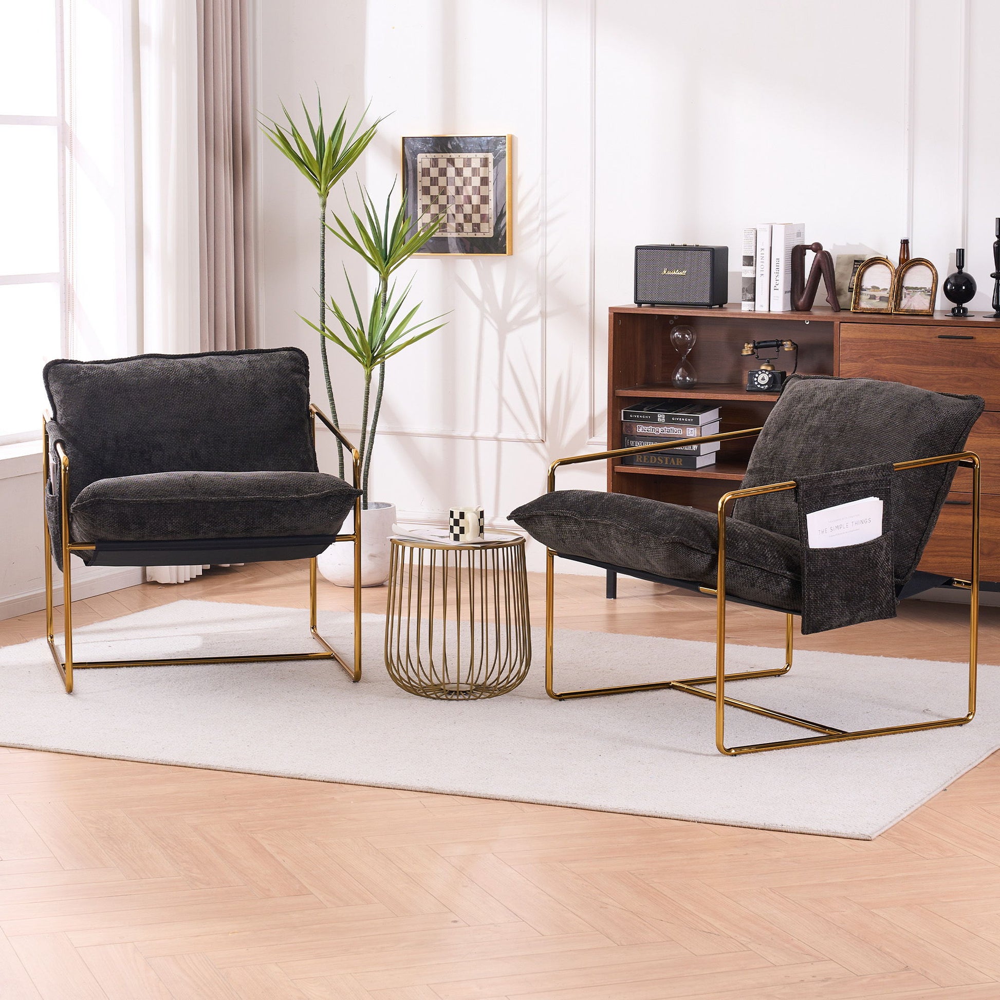 Two Upholstered Hanging Armchairs with gold-plated metal frames, crushed foam cushions, and skin-friendly woven fabric sit on a light rug by a round coffee table. Sunlight streams in, highlighting their modern design. A plant and sideboard are in the background.