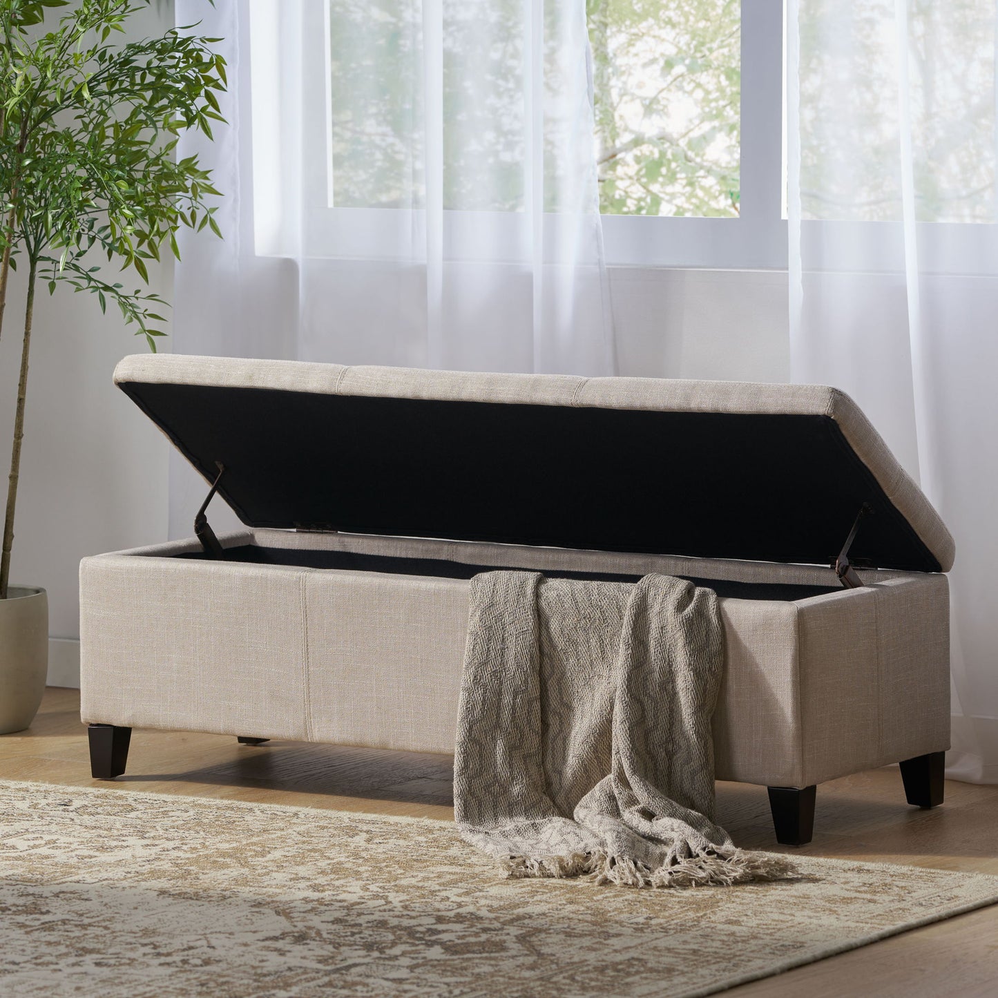 The Glouster Contemporary Storage Ottoman sits open, displaying a gray knit throw draped over the side. It's placed on a patterned rug near a window with sheer white curtains and a potted plant nearby.