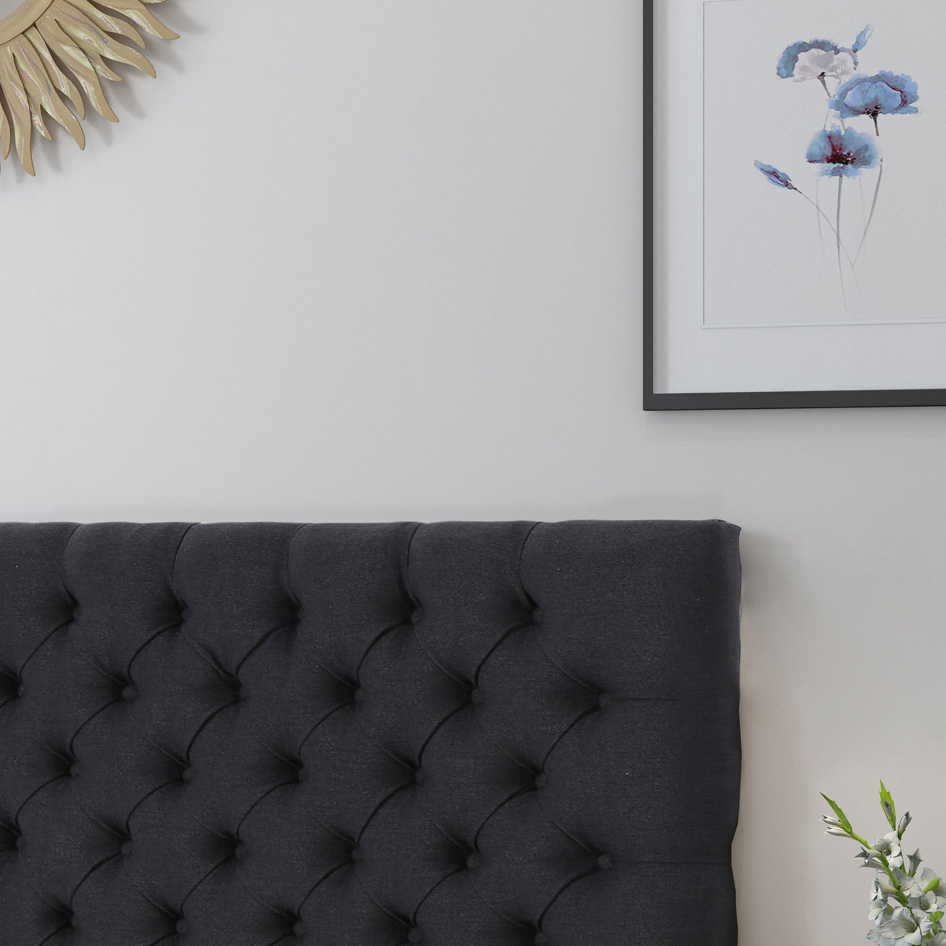 A bedroom corner featuring the Annecy - Contemporary Upholstered Headboard, a framed floral painting, part of a sunburst mirror on a light gray wall, and a small vase of white flowers.