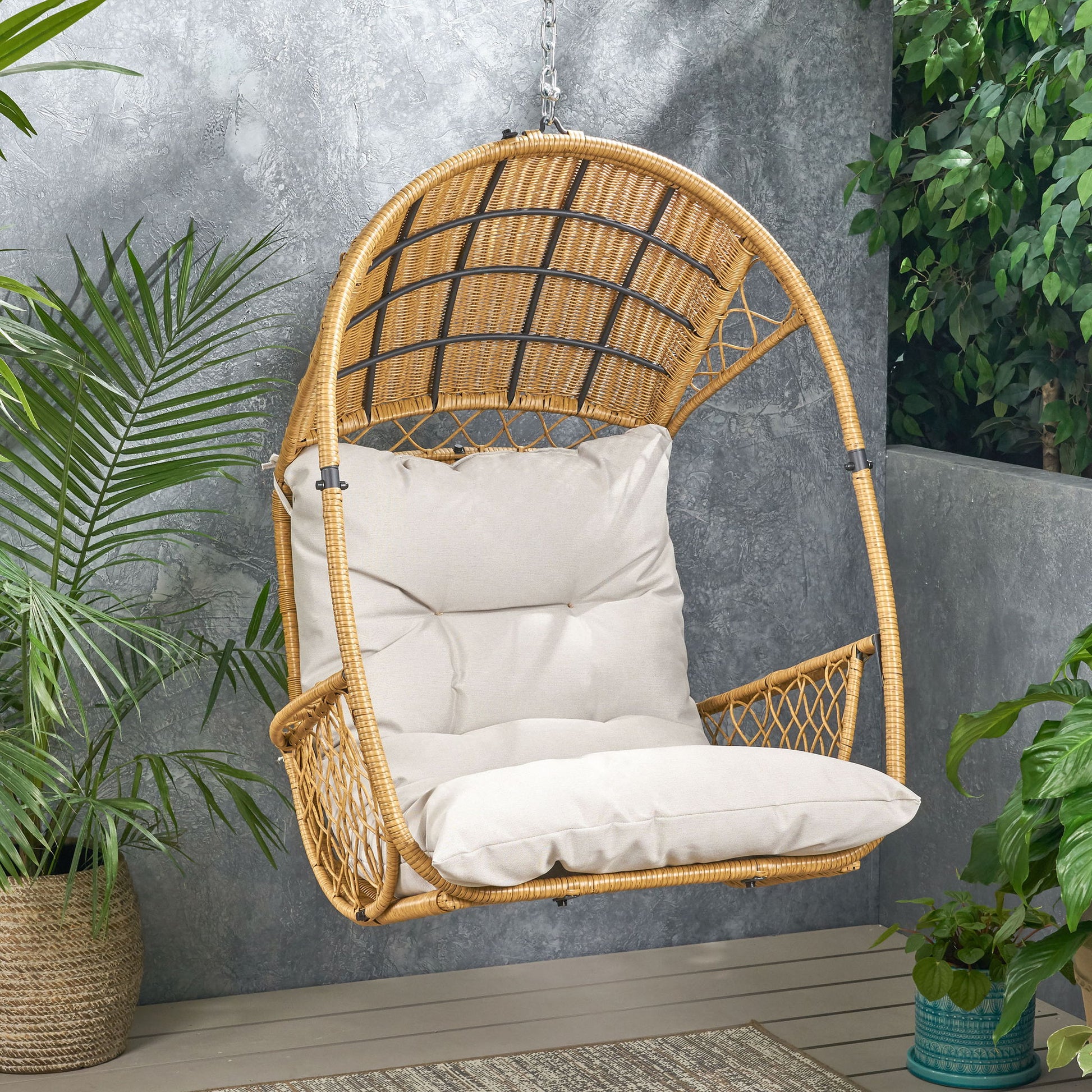 The Malia - Wicker Hanging Chair With Cushion hangs from a chain in an outdoor patio, surrounded by green potted plants and grey walls.