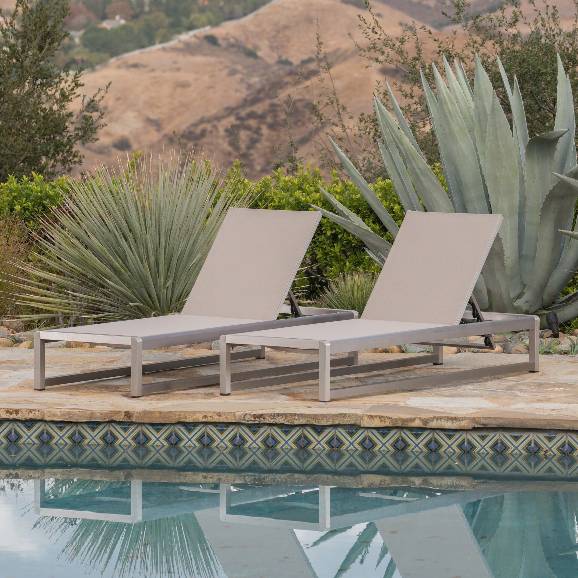 Two Outdoor Patio Chaise Lounges with 5 Reclining Positions sit poolside on a stone patio, surrounded by agave plants and greenery, with hills and trees in the background beneath a clear sky.