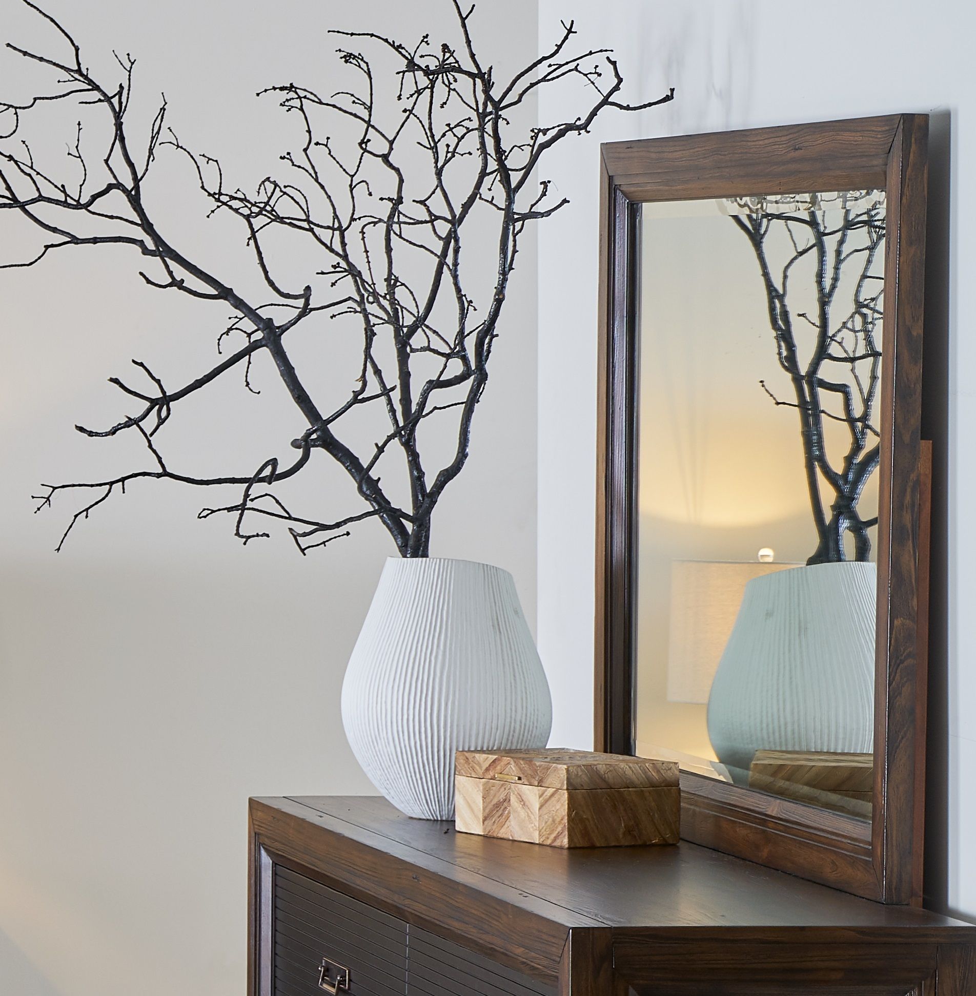 A white textured vase with black branches is on a wooden dresser beside a box, with the Couch.com Branson Mirror Rustic Buckeye in a gray finish reflecting a lamp and part of the room.