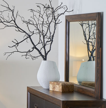A white textured vase with black branches is on a wooden dresser beside a box, with the Couch.com Branson Mirror Rustic Buckeye in a gray finish reflecting a lamp and part of the room.