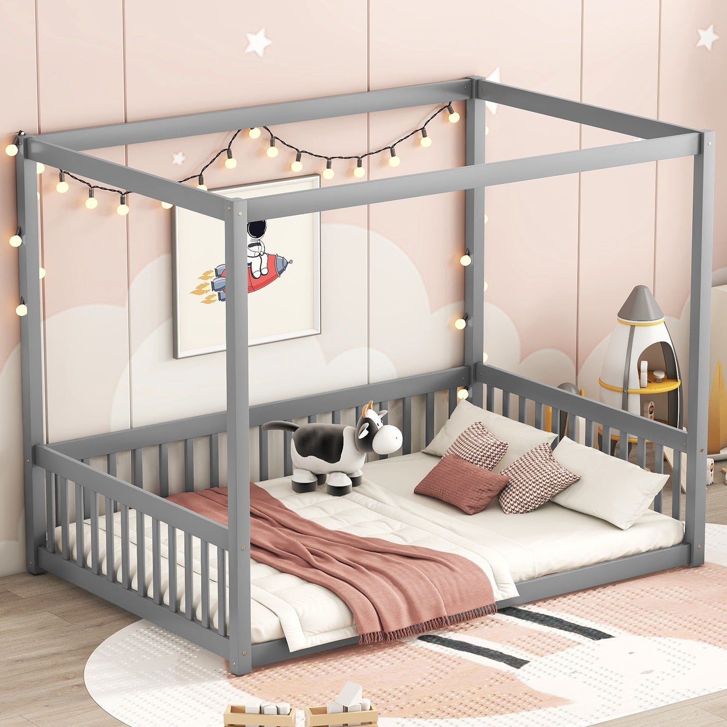 A Canopy Frame Floor Bed With Fence, Guardrails is in a children's room with pastel pink walls, a moon mural, playful decor, string lights, pillows, a pink blanket, and a stuffed cow.