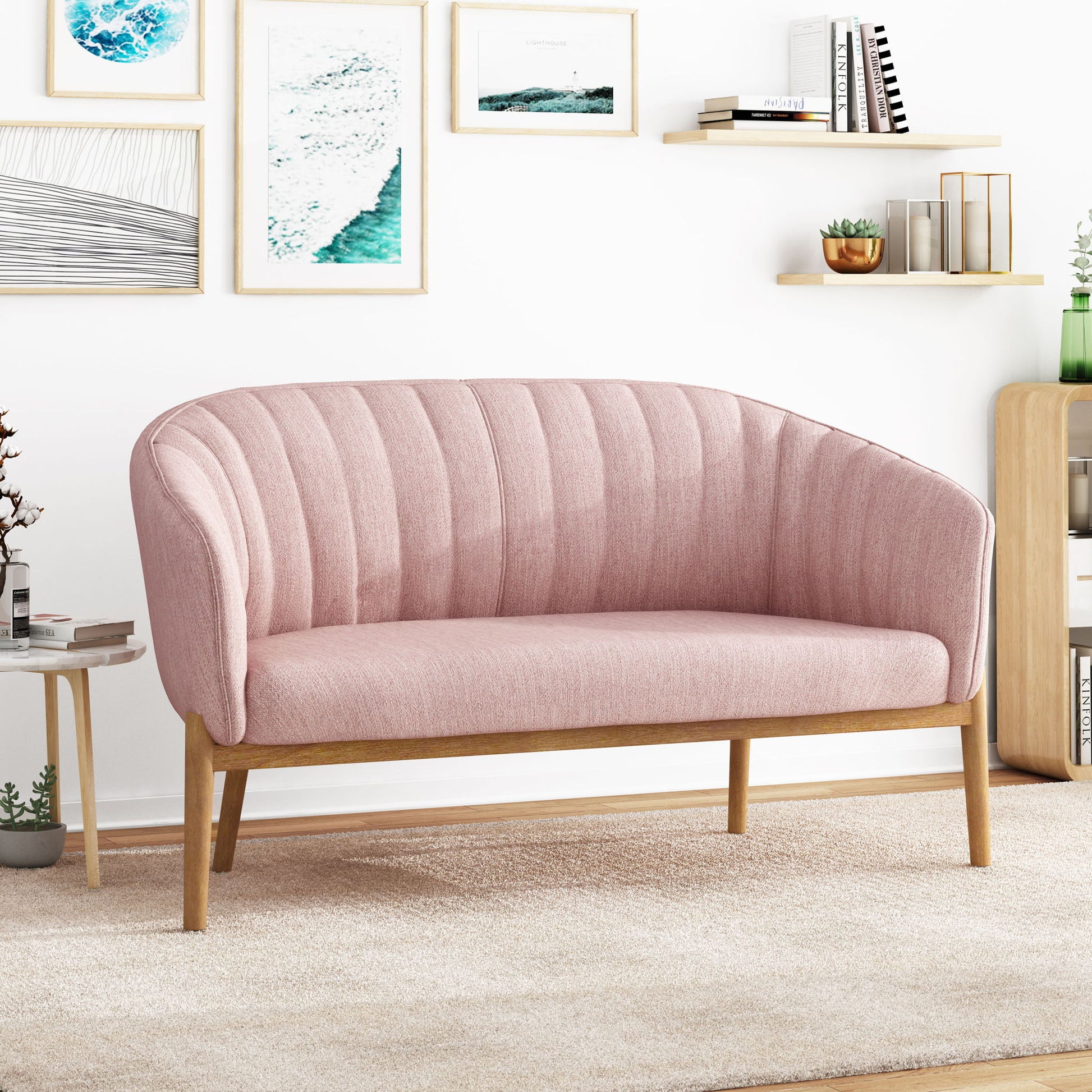A modern living room with the Mid Century Modern Loveseat With Channel Stitched Backrest in pink, wooden legs, framed art, potted plants, a small side table, a bookshelf, and a beige carpet.