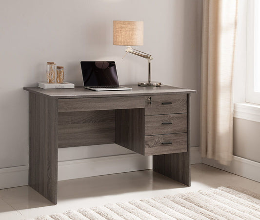 The Executive Office Desk, Lockable 3 Drawer Home Office Desk in Distressed Gray sits by a window with beige curtains and a cream rug, topped with a closed laptop, decorative jar, notebook, and silver desk lamp with beige shade.