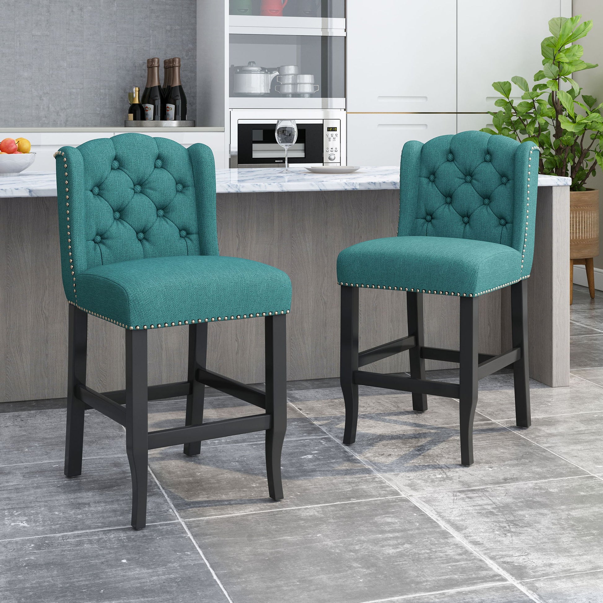 A set of Vienna Fabric Tufted Counter Stools with nailhead accents, featuring teal upholstery and black wooden legs, sits at a marble-topped kitchen island in a modern space with white cabinetry and a potted plant in the background.