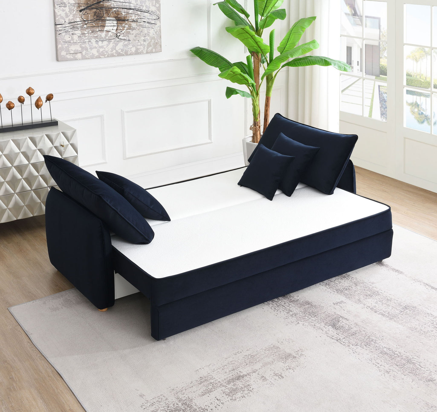 A modern living room showcases the Irina Pull Out Sleep With Storage daybed in black and white with matching pillows, a large indoor plant, abstract wall art, and big windows flooding a neutral area rug with natural light.