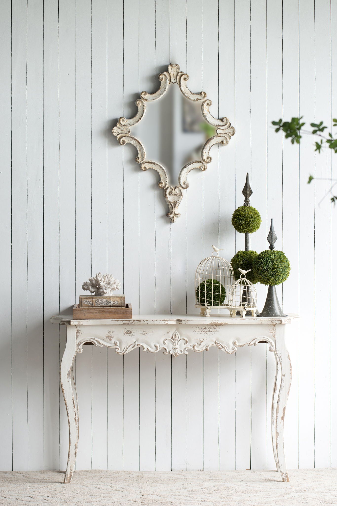 A Slimline wash Console Table, White Washed, stands against a white wood-paneled wall, topped with books, a coral sculpture, topiary plants, and birdcages. Above the table hangs an ornate decorative mirror. Perfect for hallway or living room.
