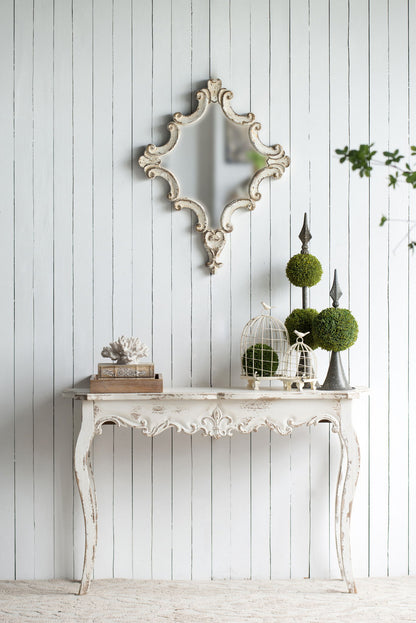 A Slimline wash Console Table, White Washed, stands against a white wood-paneled wall, topped with books, a coral sculpture, topiary plants, and birdcages. Above the table hangs an ornate decorative mirror. Perfect for hallway or living room.