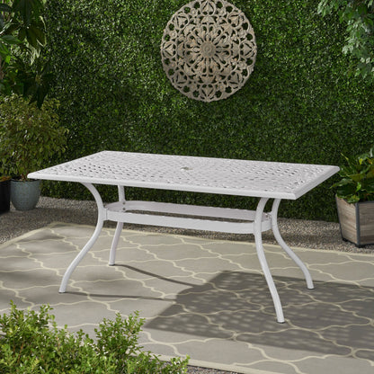 The Celestine Outdoor Cast Aluminum Dining Table with a floral lattice design and umbrella holder stands on a patterned rug in a garden with green foliage, potted plants, and circular wall art hanging on a leafy hedge.