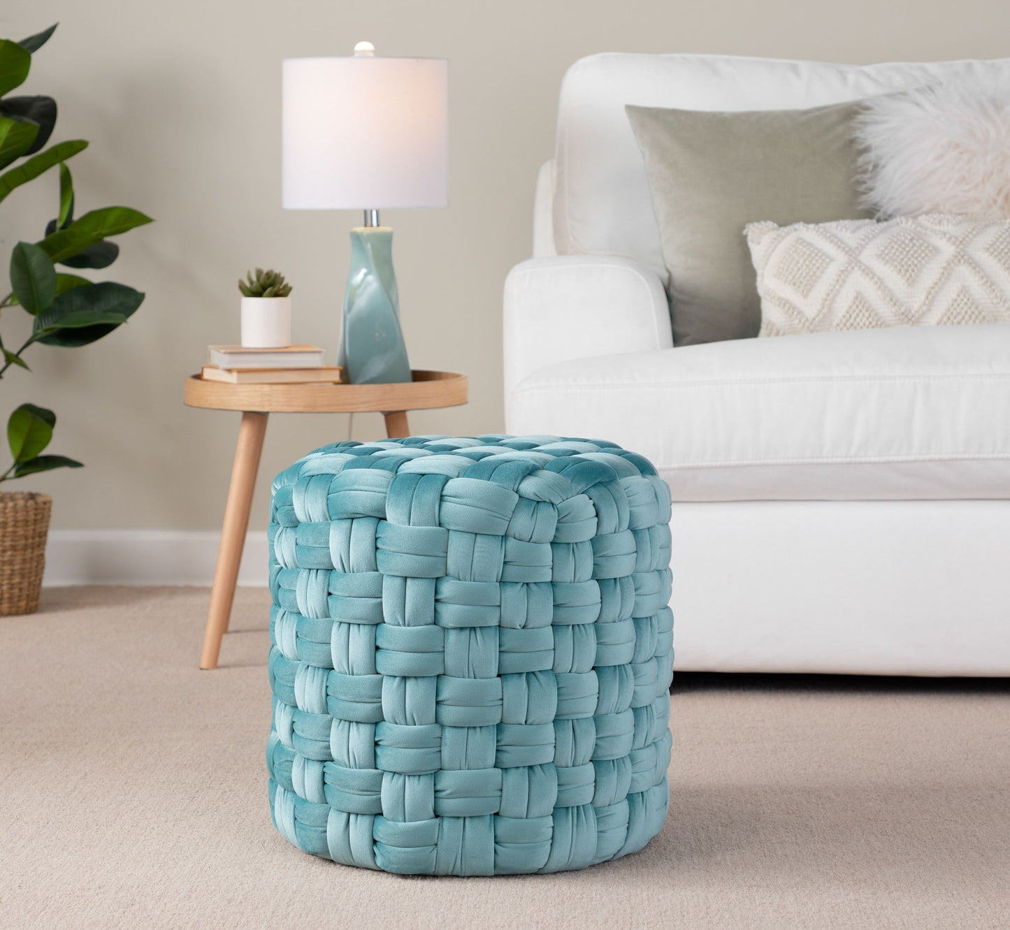 A Braided - Versatile Round Ottoman in light blue sits on a beige carpet in a living room featuring a white sofa, round wooden side table with a lamp, and a leafy plant in the background.