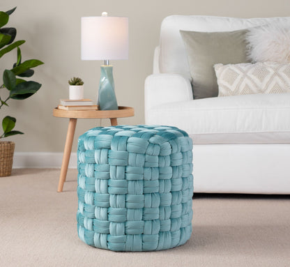 A Braided - Versatile Round Ottoman in light blue sits on a beige carpet in a living room featuring a white sofa, round wooden side table with a lamp, and a leafy plant in the background.