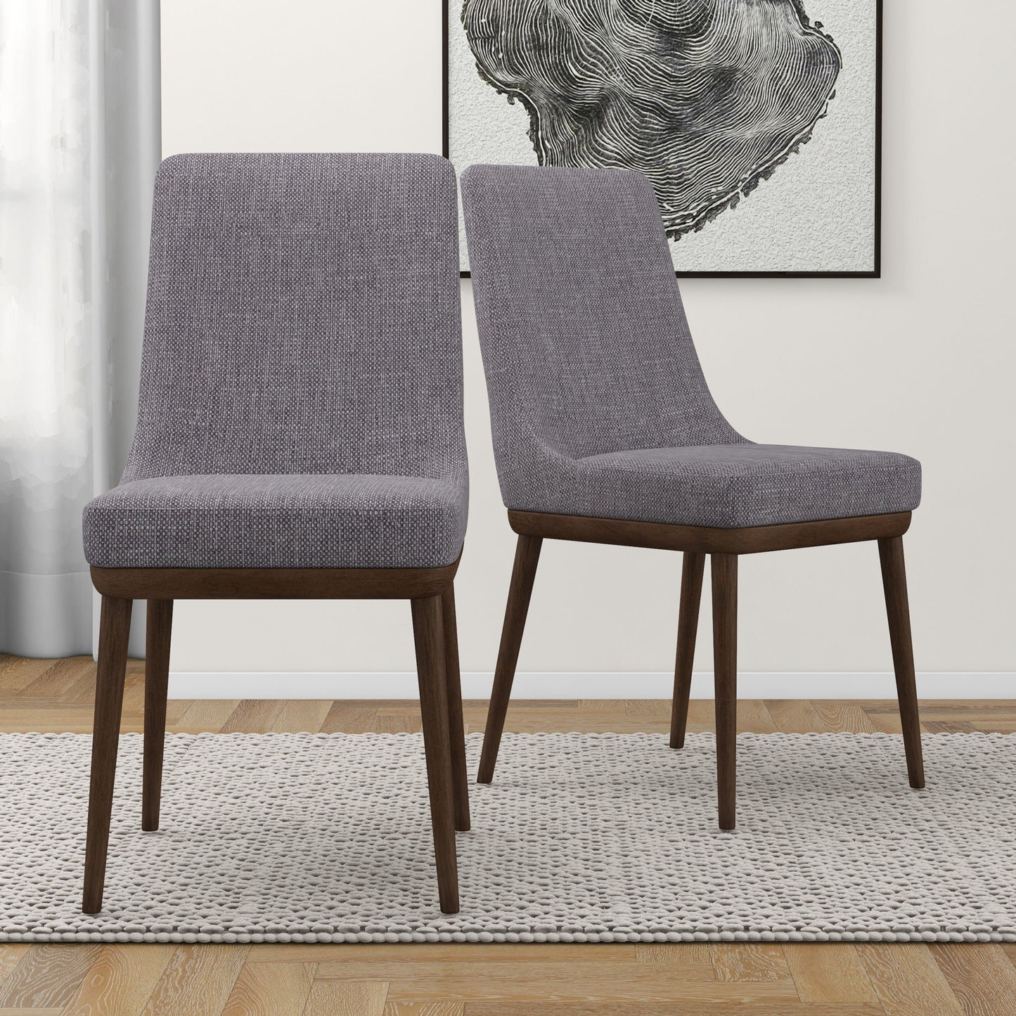 The Kate Mid-Century Modern Dining Chairs (Set of 2), featuring gray upholstery and dark wood legs, sit on a textured rug in a light-walled room with framed abstract art in the background.