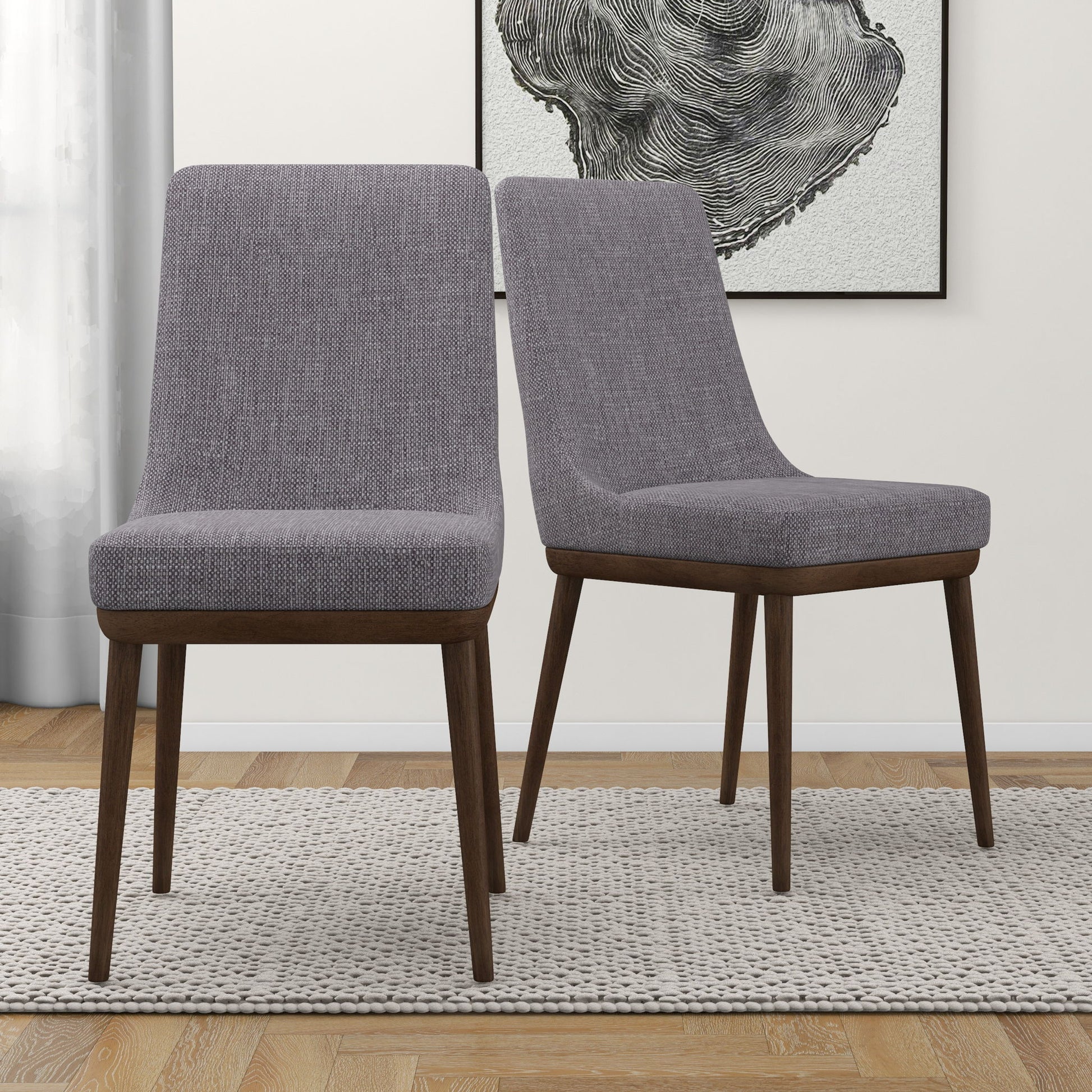 The Kate Mid-Century Modern Dining Chairs (Set of 2), featuring gray upholstery and dark wood legs, sit on a textured rug in a light-walled room with framed abstract art in the background.