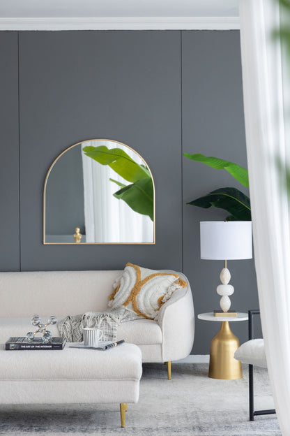 A modern living room with a gray accent wall, an Arched Mirror With Metal Frame, a white sofa with patterned pillows and throw blanket, a gold and white side table, a white lamp, and large green plant leaves.