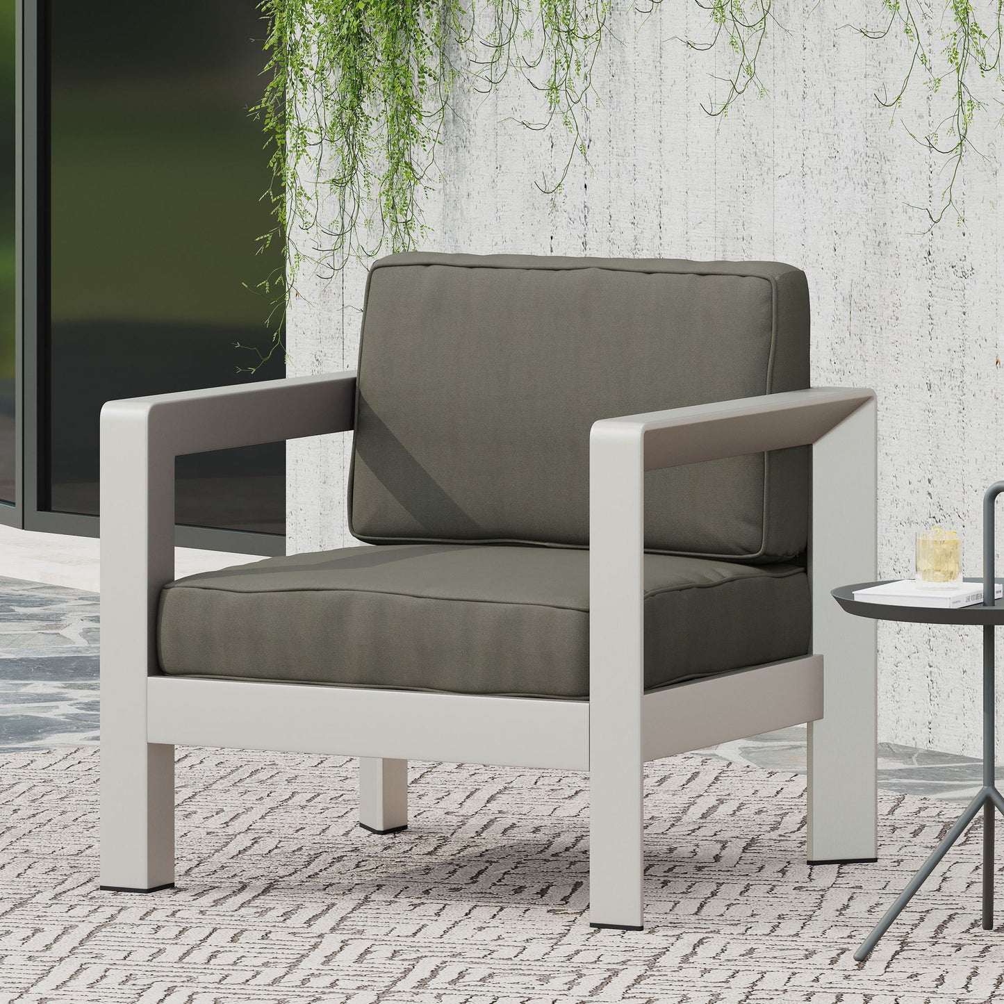 The Aviara - Luxurious Outdoor Club Chair features thick dark green cushions and a sturdy light gray frame, set on a patterned rug beside a small round side table, with greenery hanging on a concrete wall in the background.