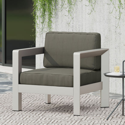 The Aviara - Luxurious Outdoor Club Chair features thick dark green cushions and a sturdy light gray frame, set on a patterned rug beside a small round side table, with greenery hanging on a concrete wall in the background.
