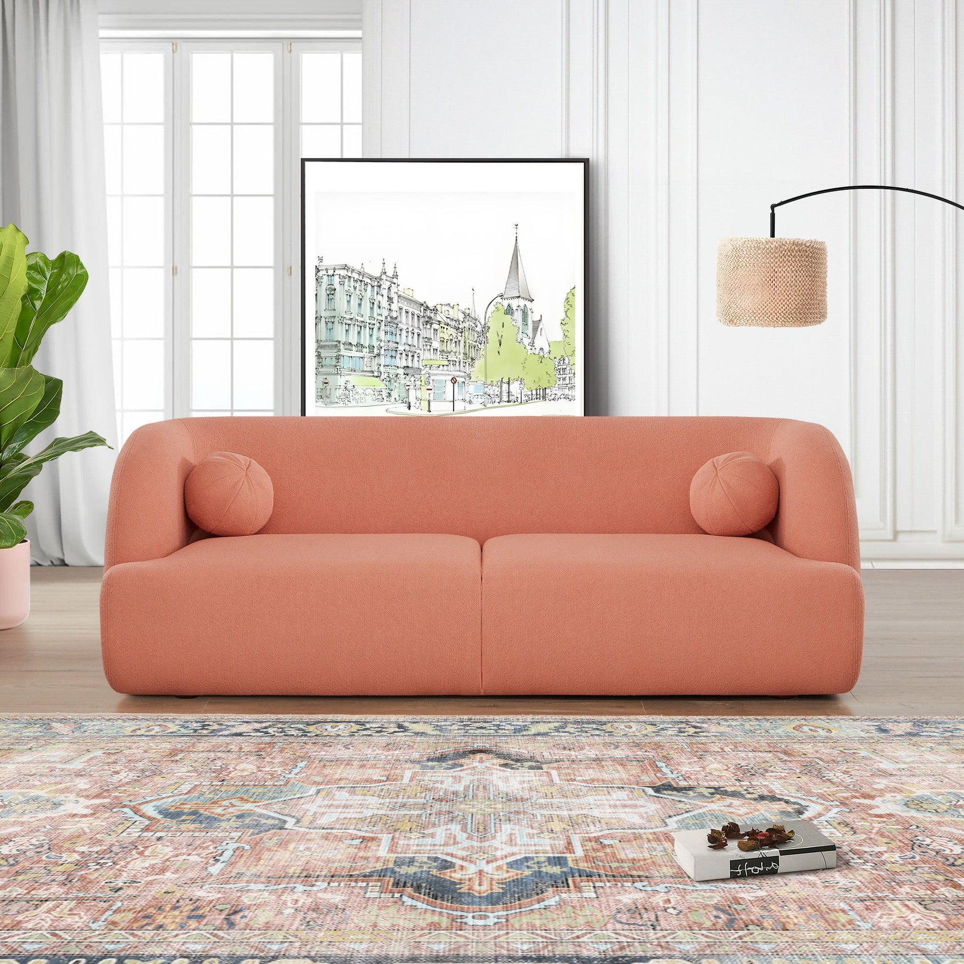 A modern living room with the Anna - French Boucle Sofa in coral, two round cushions, a large leafy plant, a patterned rug, framed cityscape artwork, and a woven floor lamp against white paneled walls and large windows.