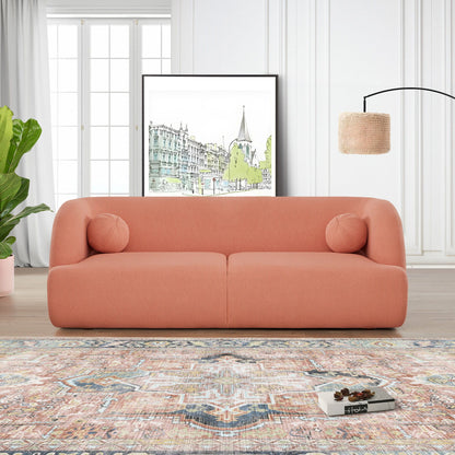 A modern living room with the Anna - French Boucle Sofa in coral, two round cushions, a large leafy plant, a patterned rug, framed cityscape artwork, and a woven floor lamp against white paneled walls and large windows.