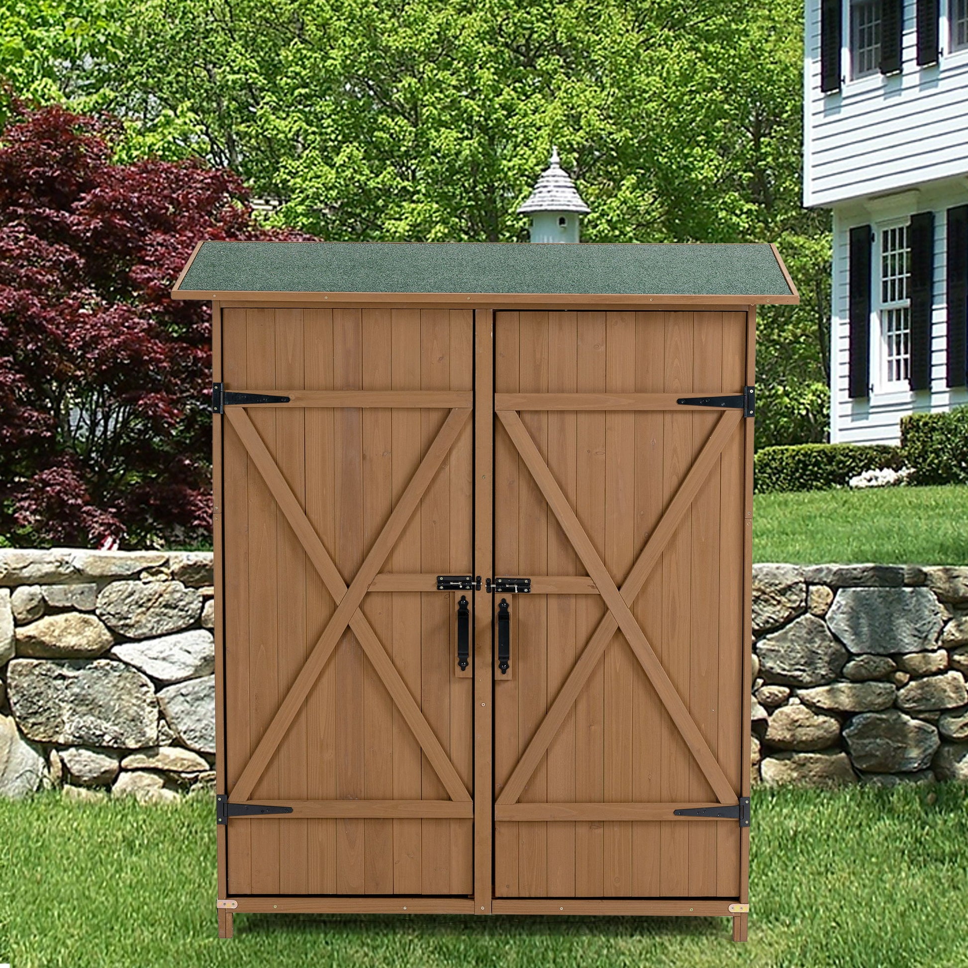 An Outdoor Storage Shed With Lockable Door, featuring detachable shelves and a pitched roof, stands on a lawn in front of a stone wall, with a house and trees in the background.