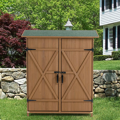 An Outdoor Storage Shed With Lockable Door, featuring detachable shelves and a pitched roof, stands on a lawn in front of a stone wall, with a house and trees in the background.